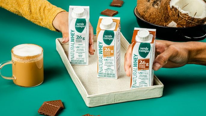 Embalagens de Natural Whey Verde Campo Lacfree com 26g de proteína organizadas em bandeja, com café servido em xícara, pedaços de chocolate e coco ao fundo, em composição de estúdio.