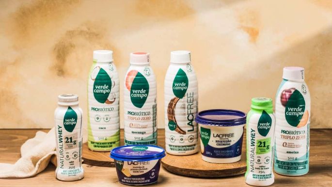 Linha completa de produtos Verde Campo Lacfree disposta sobre mesa de madeira, incluindo bebidas probióticas, Natural Whey e requeijão cremoso, em cenário rústico com fundo claro.