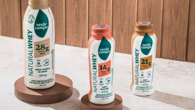Garrafas de bebida Natural Whey Verde Campo em diferentes sabores dispostas sobre pedestais em cenário moderno.