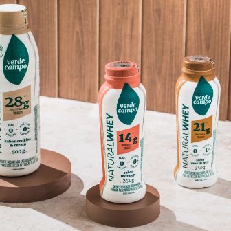 Garrafas de bebida Natural Whey Verde Campo em diferentes sabores dispostas sobre pedestais em cenário moderno.