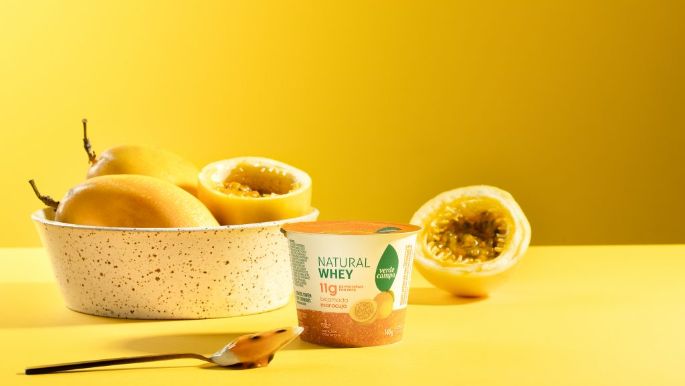 Pote de Natural Whey Verde Campo sabor maracujá disposto em estúdio, acompanhado de maracujás frescos, colher e tigela cerâmica, sobre fundo amarelo vibrante.