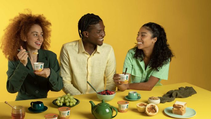 Três pessoas sentadas à mesa consumindo produtos Natural Whey Verde Campo com frutas e utensílios.