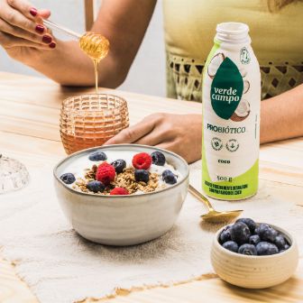 Bebida probiótica Verde Campo sabor coco ao lado de tigela com granola, frutas vermelhas e mel.