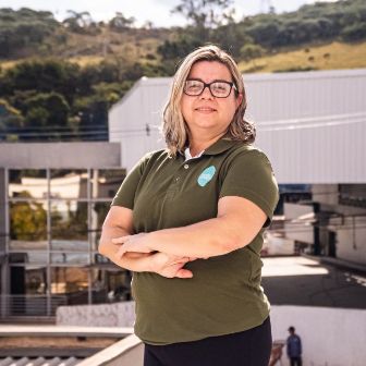 Pessoa em pé em área externa da unidade Verde Campo, posicionada em primeiro plano com braços cruzados, tendo ao fundo instalações industriais e paisagem natural.