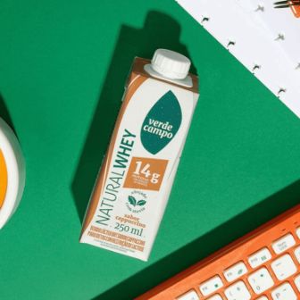 Bebida Natural Whey Verde Campo sabor cappuccino sobre mesa com relógio e objetos de escritório.