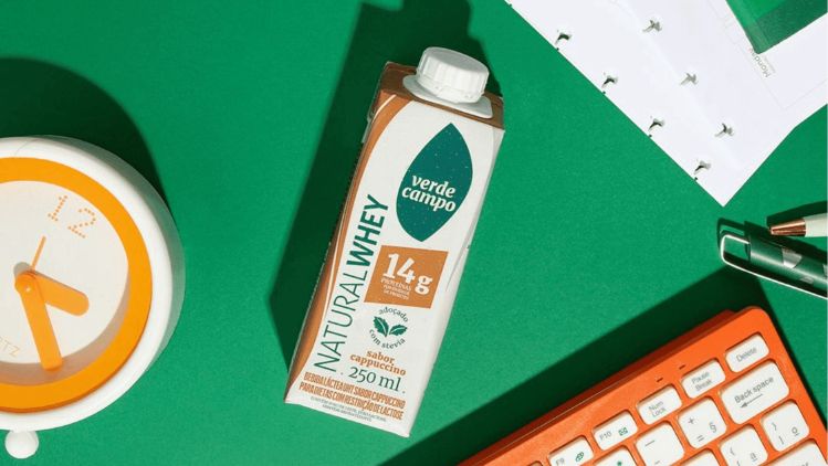 Bebida Natural Whey Verde Campo sabor cappuccino sobre mesa com relógio e objetos de escritório.