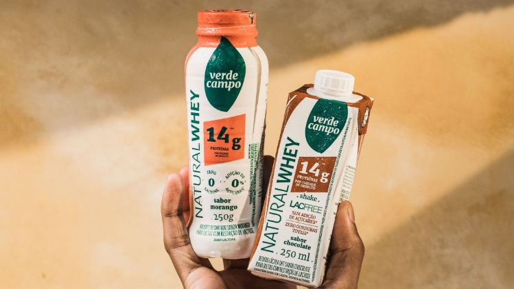 Bebidas Natural Whey Verde Campo sendo seguradas, destacando rótulos, sabores e informação de proteína.