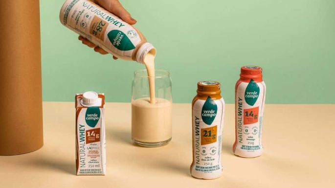 Bebidas Natural Whey Verde Campo em diferentes sabores dispostas em estúdio, com uma delas sendo servida em copo de vidro, sobre fundo verde e amarelo em composição minimalista.