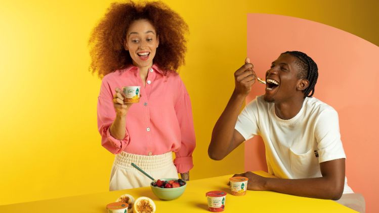 Duas pessoas à mesa consumindo produtos Verde Campo, com potes de Natural Whey, frutas frescas e fundo amarelo vibrante.