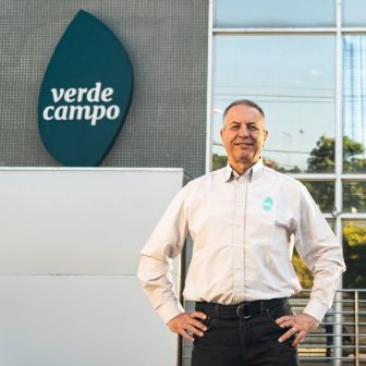 Pessoa em pé em frente à fachada da unidade Verde Campo, com logotipo da marca visível na parede e prédio envidraçado ao fundo, em ambiente corporativo externo.