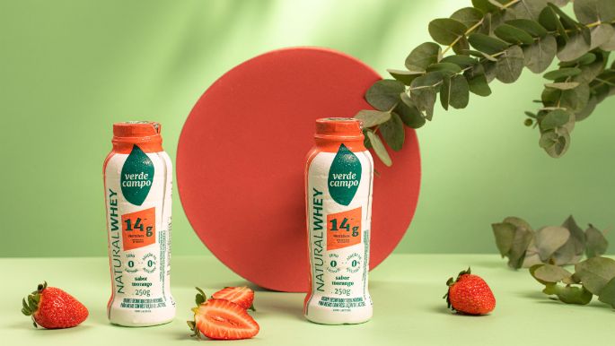 Duas garrafas de Natural Whey Verde Campo sabor morango dispostas em estúdio, acompanhadas de morangos frescos e folhas decorativas, sobre fundo verde com elemento circular vermelho ao centro.