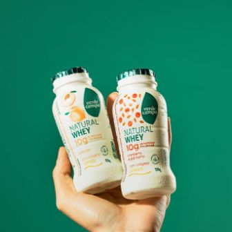Duas garrafas de Natural Whey Verde Campo com colágeno sendo seguradas em primeiro plano, destacando sabores e informações nutricionais, sobre fundo verde em estúdio.