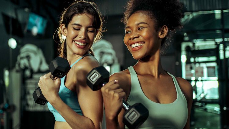 Duas pessoas levantam halteres durante treino de braço em academia.